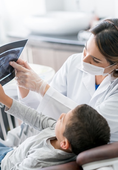 Una dentista con bata blanca y mascarilla mostrando una radiografía dental a un niño sentado en un sillón dental.