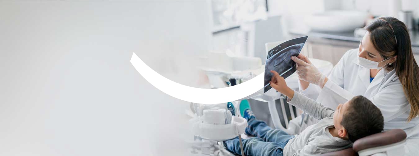 Una dentista con bata blanca y mascarilla mostrando una radiografía dental a un niño sentado en un sillón dental.