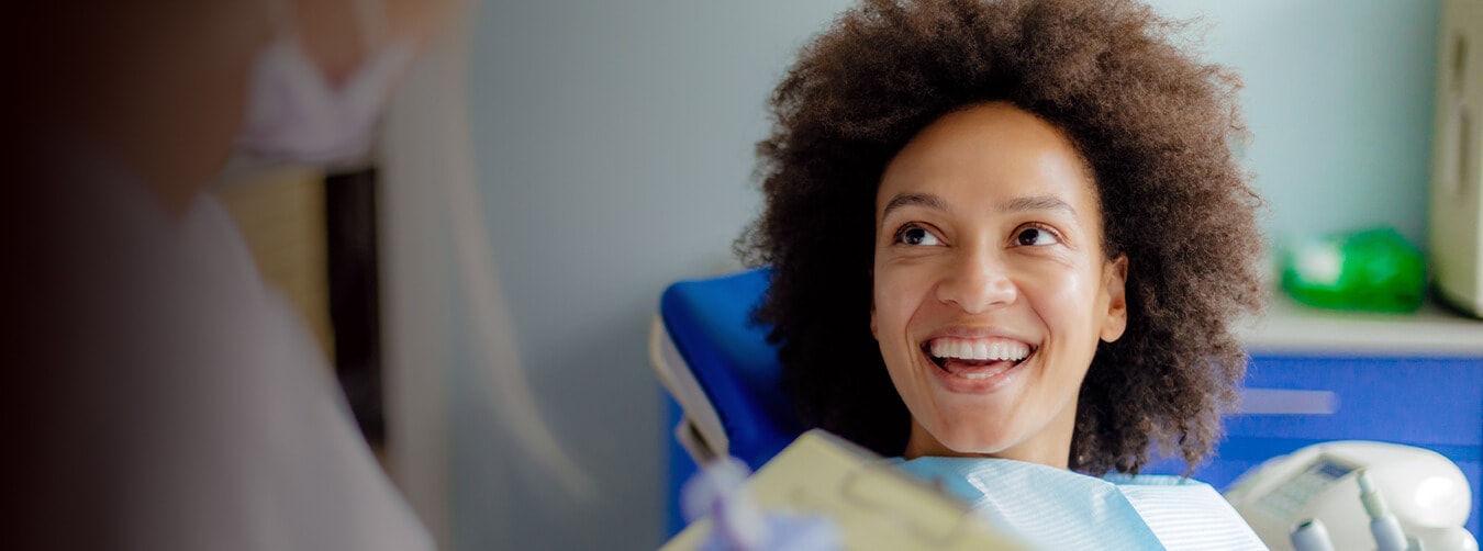 Una mujer con gran cabello rizado sonriendo y mirando al profesional dental mientras se sienta en una silla de dentista
