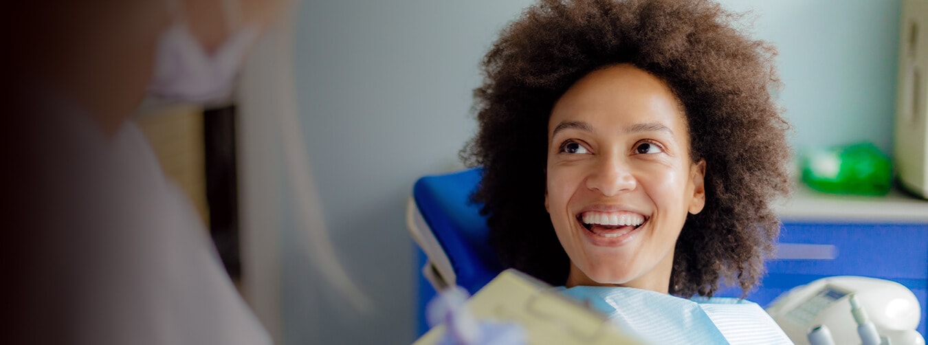 Una mujer con gran cabello rizado sonriendo y mirando al profesional dental mientras se sienta en una silla de dentista Una mujer con gran cabello rizado sonriendo y mirando al profesional dental mientras se sienta en una silla de dentista